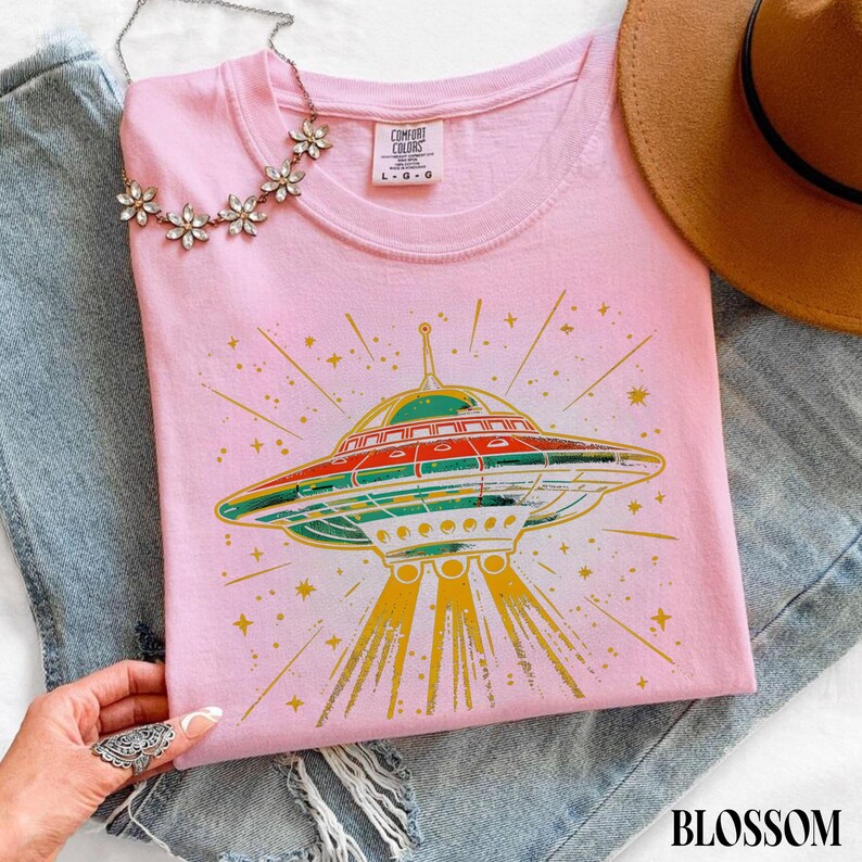 May include: A light pink t-shirt featuring a graphic of a flying saucer emitting yellow light beams and surrounded by small stars. The shirt has a crew neckline and a Comfort Colors brand label at the inner collar. The design uses red, green, and yellow.
