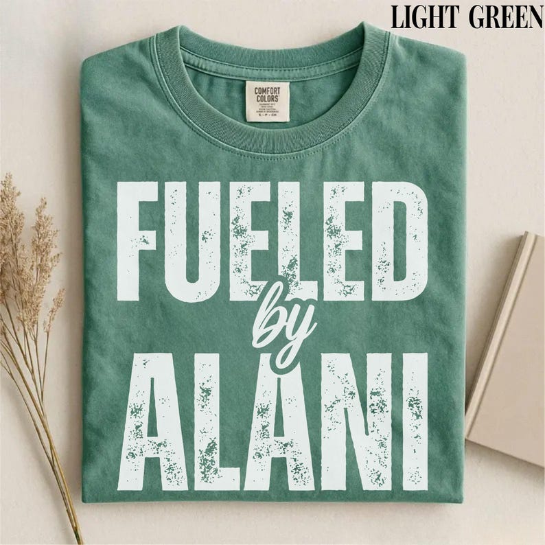 May include: Light green t-shirt with the text "FUELED by ALANI" in white block letters. The shirt has a crew neck and a tag on the collar. The shirt is folded on a light-colored surface.