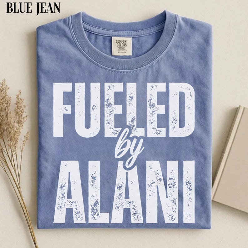 May include: Blue jean colored t-shirt with the text "FUELED by ALANI" in white, distressed lettering. The shirt has a crew neck and a Comfort Colors tag. The shirt is folded neatly on a neutral surface.