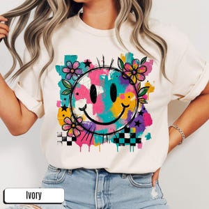 Peut inclure: T-shirt ivoire avec un grand motif de smiley coloré. Le graphisme comprend un contour noir, des fleurs, des étoiles et un motif à carreaux. Le smiley est rempli de couleurs vives comme le rose, le bleu, le jaune et le violet.