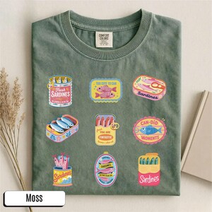 May include: A moss-green t-shirt with various sardine can designs. The graphics include phrases like "Fresh Sardines" and "Can-Did Moments". The shirt has a round neck and short sleeves. The shirt is made of a soft material.