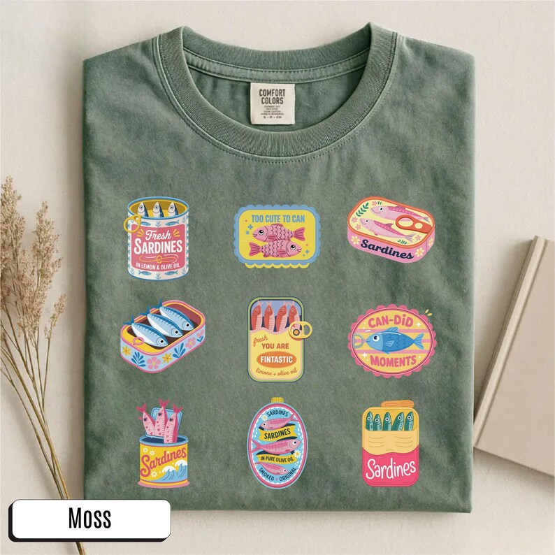 May include: A moss-green t-shirt with various sardine can designs. The graphics include phrases like "Fresh Sardines" and "Can-Did Moments". The shirt has a round neck and short sleeves. The shirt is made of a soft material.