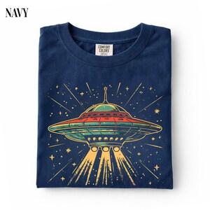 May include: A navy blue t-shirt featuring a graphic of a flying saucer with red, green, and yellow accents. The design includes a central UFO emitting light beams downward, surrounded by stars and lines. The inner neck label reads Comfort Colors.