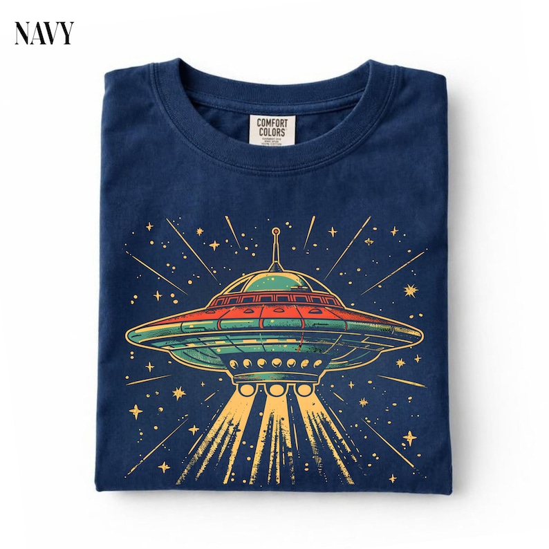 May include: A navy blue t-shirt featuring a graphic of a flying saucer with red, green, and yellow accents. The design includes a central UFO emitting light beams downward, surrounded by stars and lines. The inner neck label reads Comfort Colors.