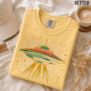 May include: A butter yellow crew neck t-shirt featuring a graphic print of a flying saucer emitting a beam of light. The design includes red, teal, and yellow colors with star accents. The shirt has a Comfort Colors brand label at the neckline.