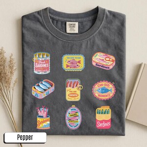 May include: Dark grey t-shirt with sardine graphics. The shirt displays various sardine can and box designs with text such as "Fresh Sardines" and "Too Cute to Can". The shirt is made of a soft material.