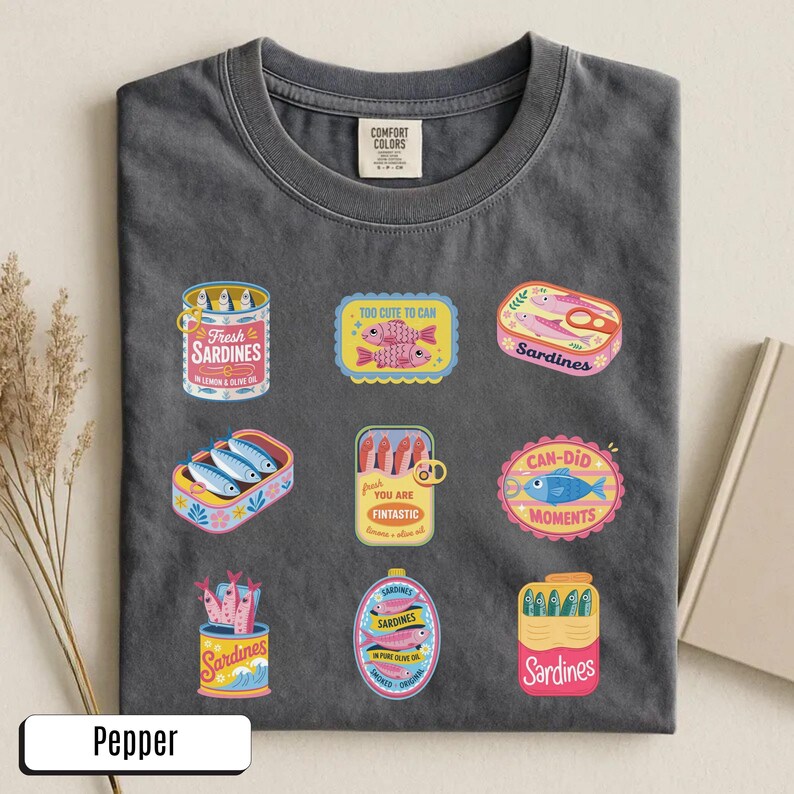 May include: Dark grey t-shirt with sardine graphics. The shirt displays various sardine can and box designs with text such as "Fresh Sardines" and "Too Cute to Can". The shirt is made of a soft material.