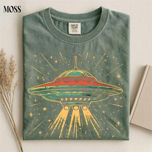 May include: A sage green crew neck t-shirt featuring a graphic of a flying saucer emitting light beams. The design includes a red and teal UFO surrounded by yellow stars and radiating lines. The inner neck label reads Comfort Colors.