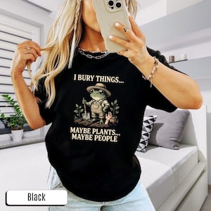 May include: Black t-shirt featuring a frog character in a hat and overalls, holding a shovel. The text on the shirt says "I bury things... Maybe plants... Maybe people."