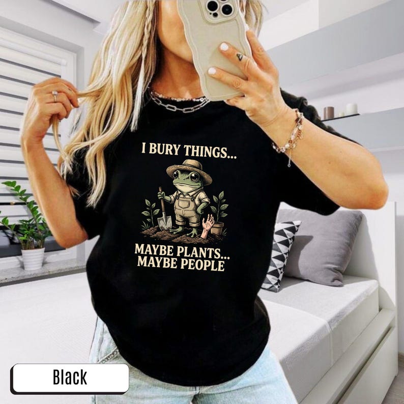 May include: Black t-shirt featuring a frog character in a hat and overalls, holding a shovel. The text on the shirt says "I bury things... Maybe plants... Maybe people."