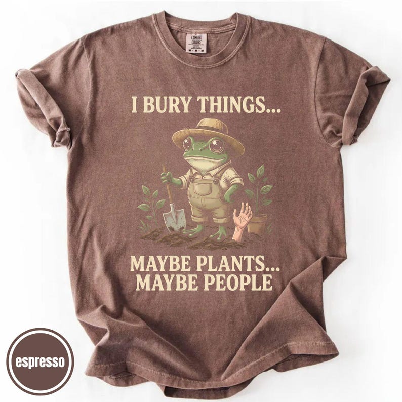 May include: Espresso-coloured t-shirt featuring a frog in a hat and overalls, holding a shovel. The text on the shirt reads "I BURY THINGS... MAYBE PLANTS... MAYBE PEOPLE."