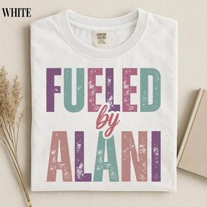 May include: White t-shirt with the text "FUELED by ALANI" in a distressed, multi-colored font. The shirt has a crew neck and a tag that reads "Comfort Colors". The shirt is folded and appears to be made of a soft material.