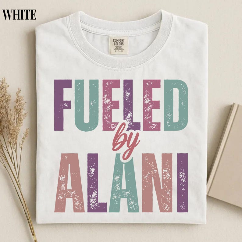 May include: White t-shirt with the text "FUELED by ALANI" in a distressed, multi-colored font. The shirt has a crew neck and a tag that reads "Comfort Colors". The shirt is folded and appears to be made of a soft material.