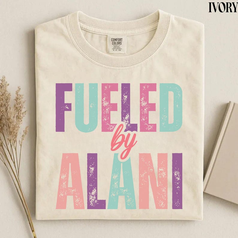 May include: Ivory-colored t-shirt with the text "FUELED by ALANI" in purple, teal, and pink block letters. The word "by" is in a cursive pink font. The shirt is folded and has a Comfort Colors tag.