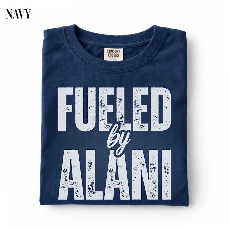 May include: Navy blue t-shirt with the text "FUELED by ALANI" in distressed white lettering. The shirt has a crew neck and a Comfort Colors tag. The word "NAVY" is in the top left corner.