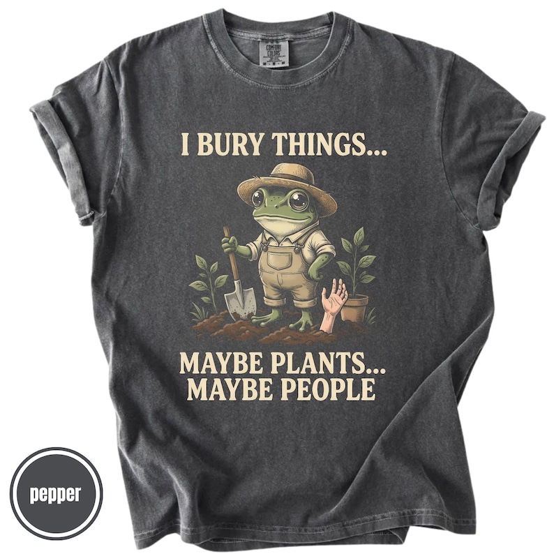 May include: Dark grey t-shirt with the text "I BURY THINGS... MAYBE PLANTS... MAYBE PEOPLE." The graphic shows a frog in overalls and a hat, holding a shovel. A hand emerges from the soil near a potted plant.
