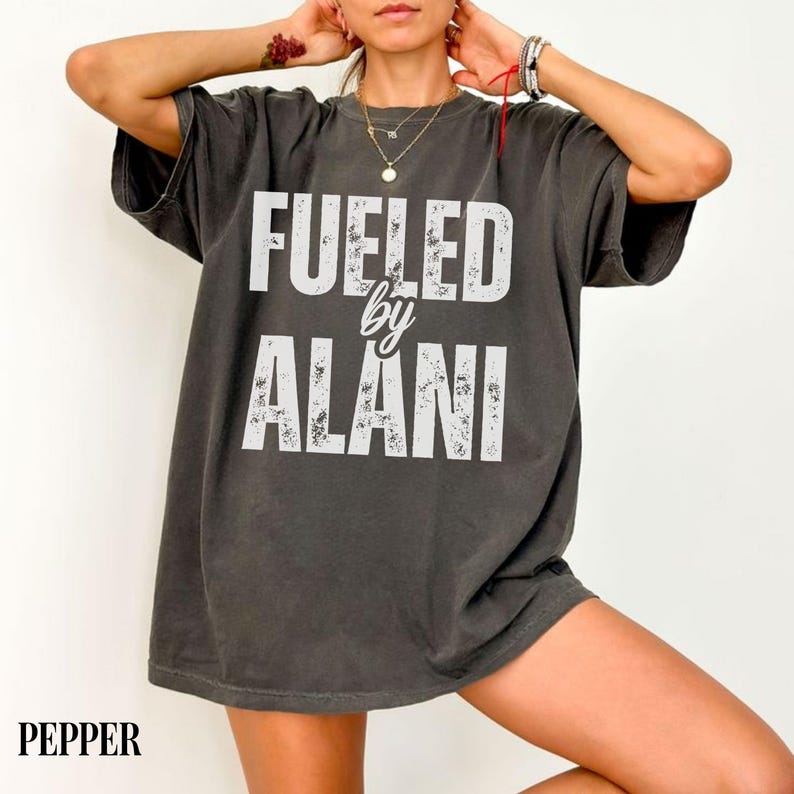 May include: Dark gray t-shirt with the text "FUELED by ALANI" in white, distressed lettering. The shirt has short sleeves and a relaxed fit. The word "PEPPER" is printed at the bottom left corner.