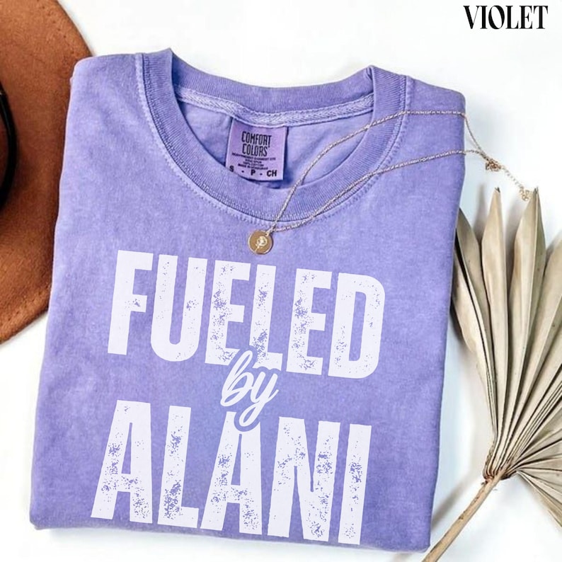 May include: A violet Comfort Colors t-shirt with the text "Fueled by Alani" in white. A gold necklace with a pendant rests on the shirt. The shirt is folded, and a brown hat and dried plant are in the background.
