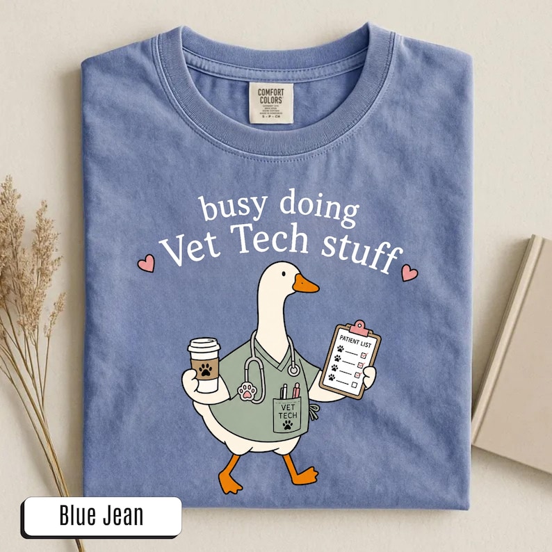 May include: A blue jean colored t-shirt featuring a graphic of a white goose dressed as a veterinary technician. The goose wears a sage green scrub top with a stethoscope, holds a coffee cup and a patient list clipboard, and has the text busy doing Vet Tech stuff printed above it.