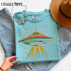 May include: A light blue t-shirt featuring a graphic of a flying saucer emitting yellow light beams and surrounded by stars. The shirt has a Comfort Colors label at the neck. A silver-toned floral necklace and a brown hat are also visible.