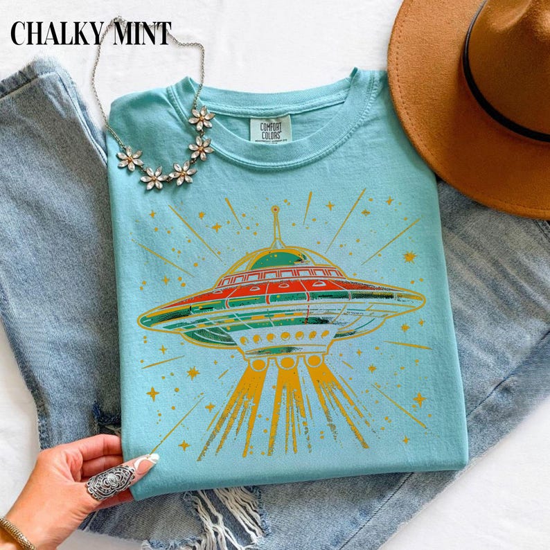 May include: A light blue t-shirt featuring a graphic of a flying saucer emitting yellow light beams and surrounded by stars. The shirt has a Comfort Colors label at the neck. A silver-toned floral necklace and a brown hat are also visible.