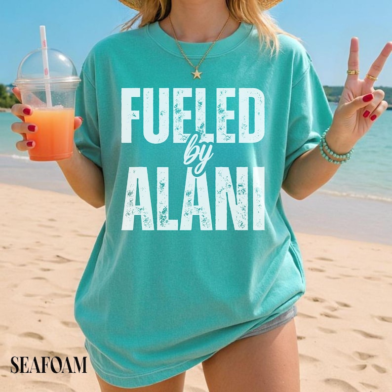 May include: Seafoam green t-shirt with the text "Fueled by Alani" in white block letters. The shirt is a relaxed fit and is worn on a beach. A clear plastic cup with a straw and orange drink is held in the left hand.