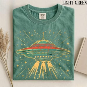 May include: A light green crew neck t-shirt featuring a graphic of a flying saucer emitting light beams. The design includes orange and yellow accents with star shapes. The shirt has a Comfort Colors brand label on the inside of the collar.