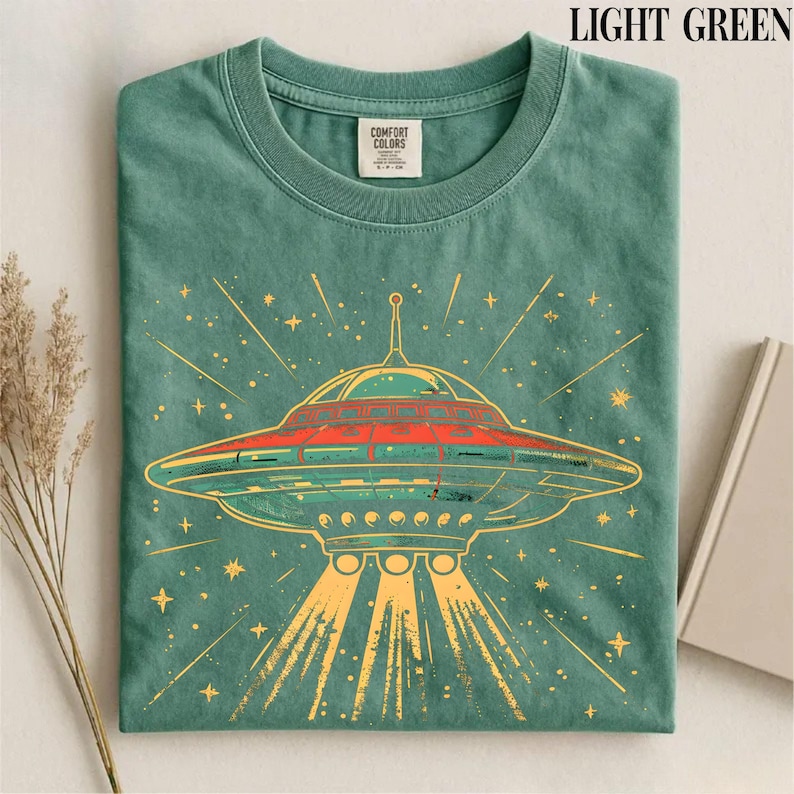 May include: A light green crew neck t-shirt featuring a graphic of a flying saucer emitting light beams. The design includes orange and yellow accents with star shapes. The shirt has a Comfort Colors brand label on the inside of the collar.