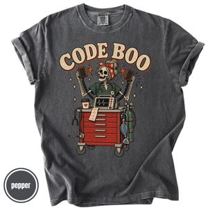 May include: A charcoal gray t-shirt featuring a graphic of a skeleton wearing a red bandana and scrubs, standing behind a red medical cart. The skeleton holds defibrillator paddles. The text CODE BOO is printed in large beige letters above the figure.