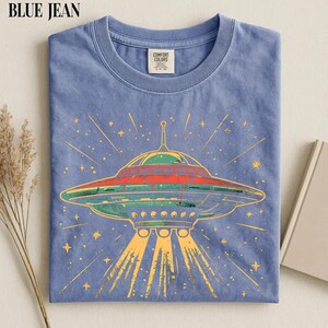 May include: A blue jean colored t-shirt featuring a graphic print of a flying saucer emitting yellow light beams. The design includes stars and lines radiating from the craft. The shirt has a crew neckline and a Comfort Colors brand label.