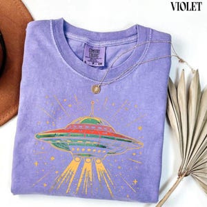 May include: A violet short-sleeve t-shirt with a graphic print of a flying saucer emitting light beams. The shirt features a Comfort Colors label at the inner neckline and is accented with a thin gold-toned chain necklace resting on the fabric.