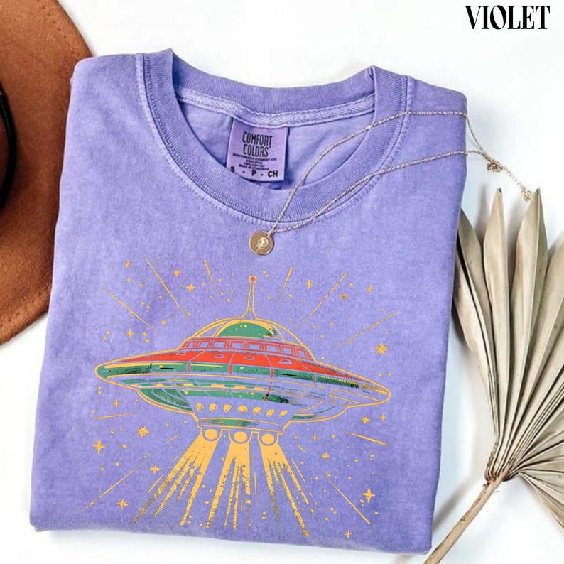 May include: A violet short-sleeve t-shirt with a graphic print of a flying saucer emitting light beams. The shirt features a Comfort Colors label at the inner neckline and is accented with a thin gold-toned chain necklace resting on the fabric.