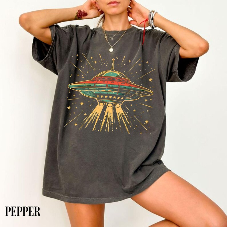 May include: A charcoal gray short-sleeve t-shirt featuring a graphic print of a flying saucer emitting yellow light beams. The design includes red and green accents on the spacecraft, surrounded by small yellow stars on a dark fabric background.