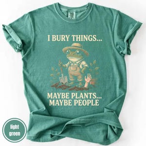 May include: Light green t-shirt with the text "I bury things... Maybe plants... Maybe people." A cartoon frog wearing overalls and a hat is holding a shovel. A hand emerges from the dirt near a potted plant.