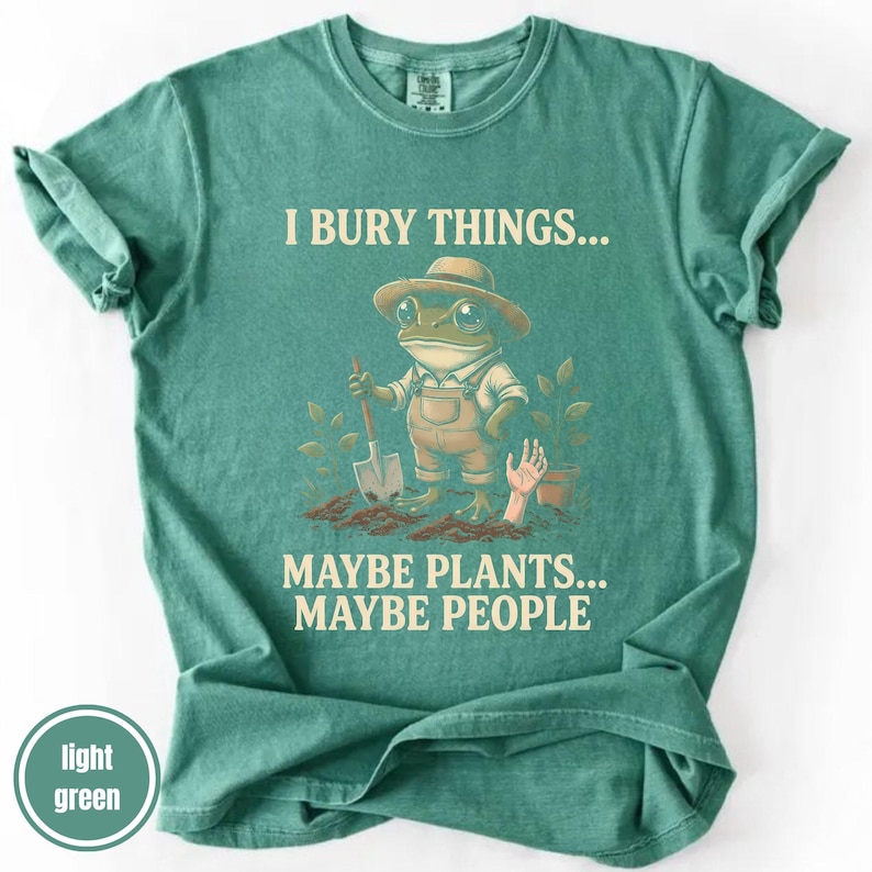 May include: Light green t-shirt with the text "I bury things... Maybe plants... Maybe people." A cartoon frog wearing overalls and a hat is holding a shovel. A hand emerges from the dirt near a potted plant.