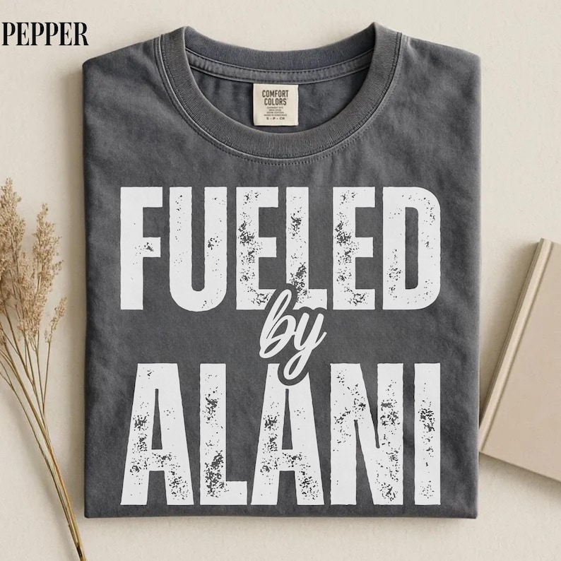May include: Dark gray t-shirt with the text "FUELED by ALANI" in distressed white lettering. The shirt has a crew neck and a Comfort Colors tag. The shirt is folded and appears to be made of soft material.