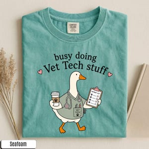 May include: A seafoam green t-shirt featuring a graphic of a white goose dressed as a veterinary technician. The goose holds a coffee cup and a patient list clipboard, wearing a stethoscope and scrubs. The text reads busy doing Vet Tech stuff.