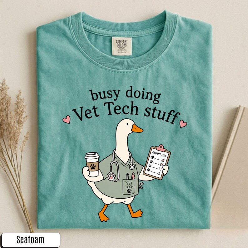 May include: A seafoam green t-shirt featuring a graphic of a white goose dressed as a veterinary technician. The goose holds a coffee cup and a patient list clipboard, wearing a stethoscope and scrubs. The text reads busy doing Vet Tech stuff.