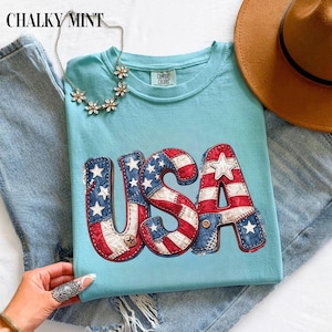 Peut inclure: Un t-shirt bleu clair avec le texte USA dans un motif de drapeau américain vieilli aux couleurs rouge, blanc et bleu. Le t-shirt a une encolure ronde et une étiquette Comfort Colors. Un collier argenté en forme de fleur est posé dessus.