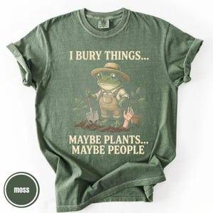 May include: Moss green t-shirt with the text "I BURY THINGS... MAYBE PLANTS... MAYBE PEOPLE." The graphic features a frog wearing overalls and a hat, holding a shovel. A hand emerges from the soil near a small pot.