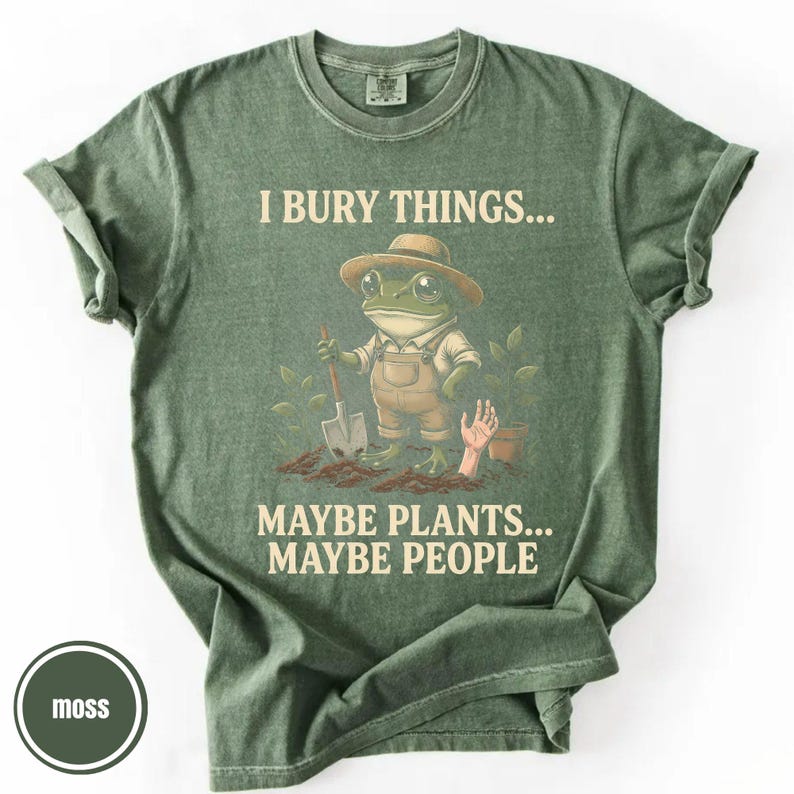 May include: Moss green t-shirt with the text "I BURY THINGS... MAYBE PLANTS... MAYBE PEOPLE." The graphic features a frog wearing overalls and a hat, holding a shovel. A hand emerges from the soil near a small pot.