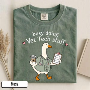 May include: A moss green t-shirt featuring a graphic of a white goose dressed as a veterinary technician. The goose holds a coffee cup and a patient list clipboard, wearing a stethoscope and scrubs. The text reads busy doing Vet Tech stuff.