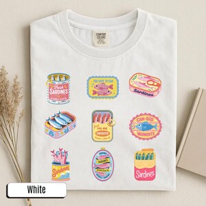 May include: White t-shirt with various sardine can illustrations. The designs include phrases such as "Fresh Sardines" and "Can-Did Moments". The shirt has a crew neck and short sleeves.