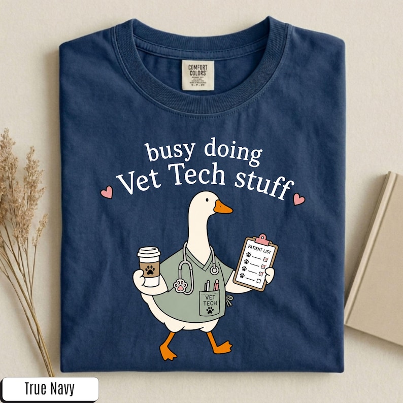 May include: A navy blue t-shirt featuring a graphic of a white goose dressed as a veterinary technician. The goose wears a light green scrub top, a stethoscope, and holds a coffee cup and a patient list clipboard. Text reads: busy doing Vet Tech stuff.