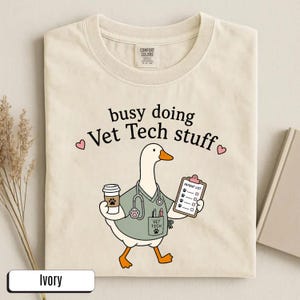 May include: An ivory t-shirt featuring a graphic of a white goose dressed in a sage green scrub top with a stethoscope, holding a coffee cup and a clipboard labeled Patient List. The text reads busy doing Vet Tech stuff with two small pink hearts.