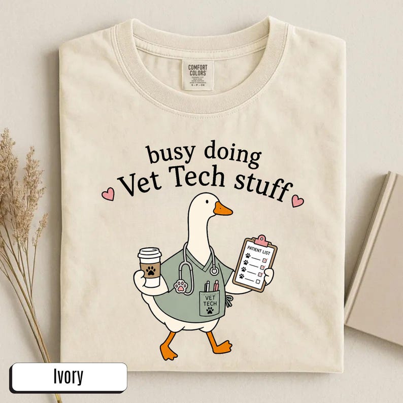 May include: An ivory t-shirt featuring a graphic of a white goose dressed in a sage green scrub top with a stethoscope, holding a coffee cup and a clipboard labeled Patient List. The text reads busy doing Vet Tech stuff with two small pink hearts.