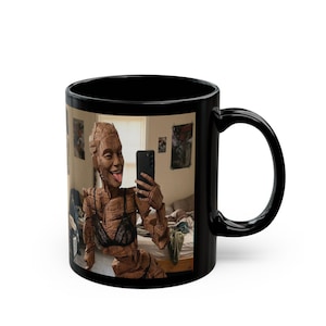 Könnte beinhalten: Schwarze Keramik-Kaffeetasse mit einem Aufdruck eines Holzskulptur-Selfies. Die Skulptur trägt einen schwarzen BH und hält ein Smartphone. Der Hintergrund zeigt ein Schlafzimmer mit Postern.