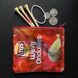 May include: A repurposed snack bag zipper pouch with golf tees, coins, and a white hair tie. The pouch is made from a Lay's Wavy Ondulées Original potato chip bag, featuring red, orange, and white text and graphics.