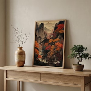 May include: A framed landscape art print depicts a mountain scene with vibrant autumn foliage in shades of red, orange, and yellow. A bonsai tree in a ceramic pot and a decorative vase with branches sit on a wooden table.