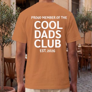 Puede incluir: Una camiseta naranja con el texto "PROUD MEMBER OF THE COOL DADS CLUB EST. 2026" impreso en blanco. La camiseta es de color liso y tiene mangas cortas. La imagen muestra la parte trasera de la camiseta.